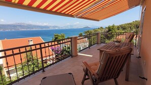 Een balkon met een houten tafel en stoelen, uitzicht op de zee, en een rood-geel gestreept zonnescherm.