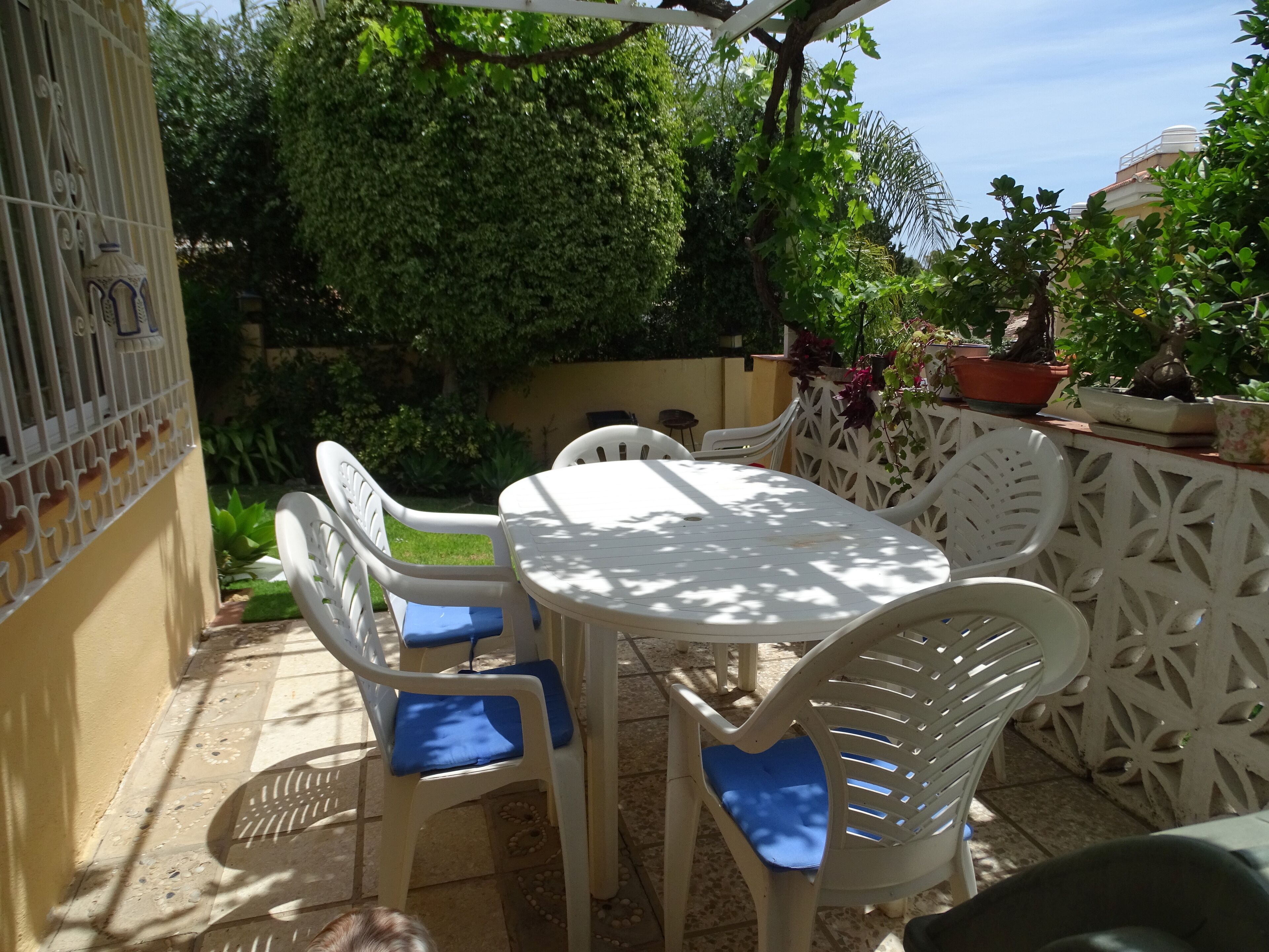 Une terrasse extérieure avec une table et des chaises blanches, entourée de verdure et de plantes en pot.