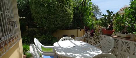Une terrasse extérieure avec une table et des chaises blanches, entourée de verdure et de plantes en pot.