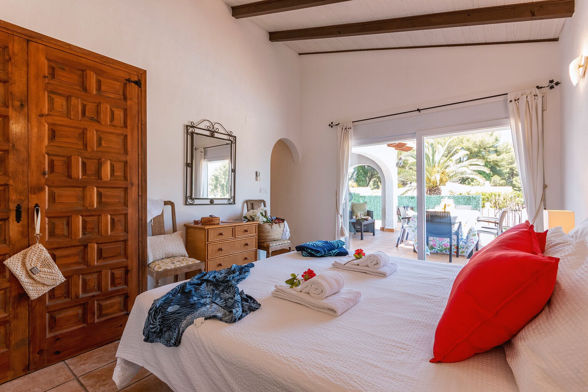 Ein Schlafzimmer mit einem großen Bett, einer Holztür, einem Schminktisch, einem Spiegel und Blick auf eine Außenveranda mit Tischen und Stühlen.