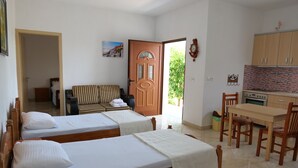 A room with two single beds, a wooden sofa, a dining table with chairs, a kitchen area, and a door leading outside. - Pellumb Apartments (Ksamil)