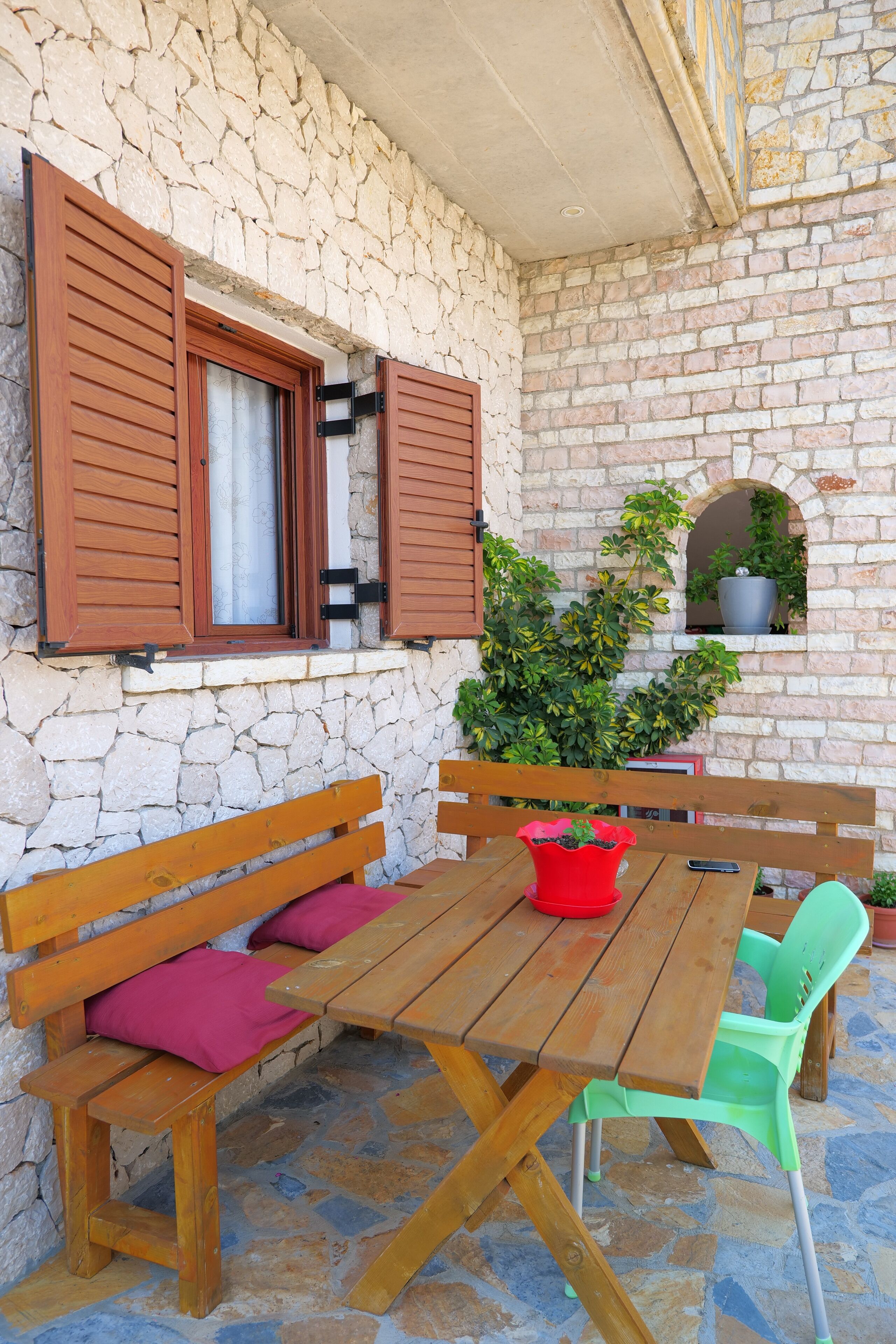 A stone wall with wooden shutters, a wooden table with benches, and potted plants.