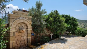 A stone archway leading to a courtyard with a stone floor, surrounded by greenery and a clear sky. - Pellumb Apartments (Ksamil)
