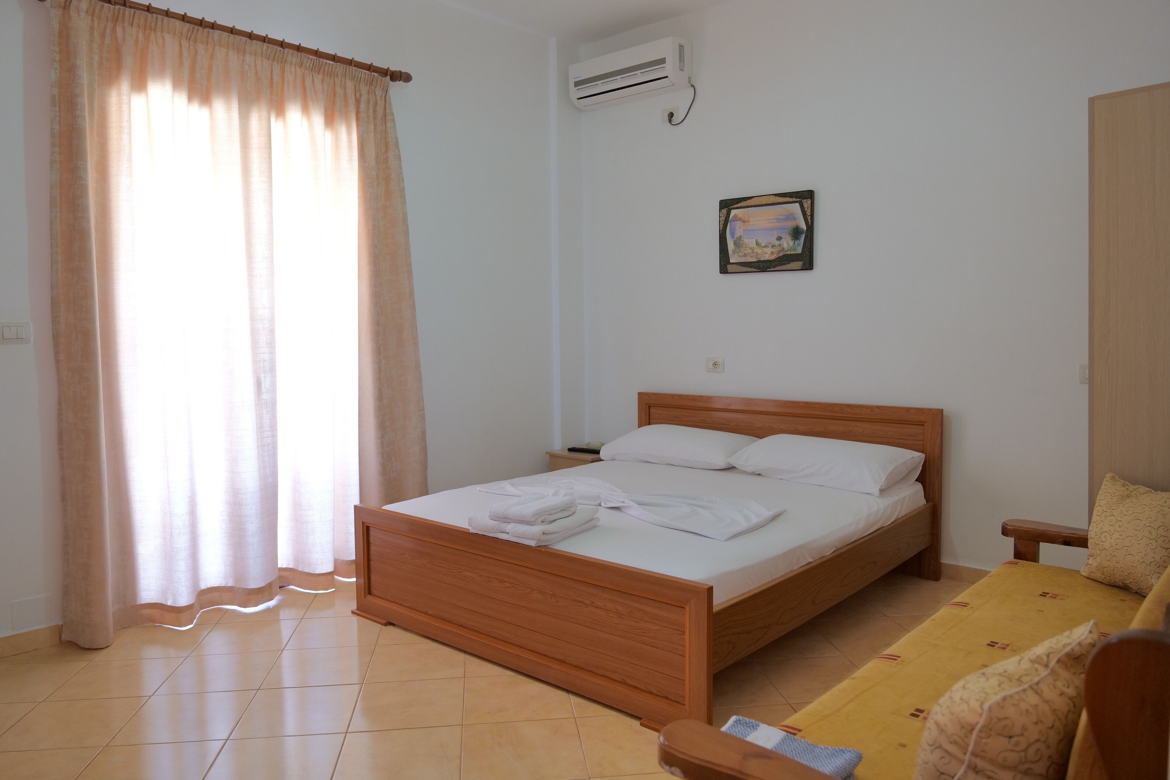 A neatly arranged bedroom with a wooden bed, a bedside table, a yellow sofa, and a wall-mounted air conditioner.