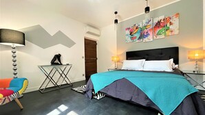 Ein modernes Hotelzimmer mit einem Bett, zwei Nachttischlampen, einem zebra-gemusterten Teppich, einem bunten Sessel, einem kleinen Tisch mit einer Skulptur und abstrakter Kunst an der Wand.