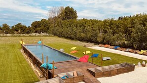 Ein Poolbereich mit Holzterrasse, bunten Kissen und einem Rasen.