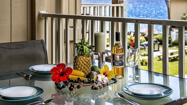 Mesa posta para refeição com pratos, talheres, uma garrafa de vinho e uma cesta de frutas numa varanda com vista para o oceano.