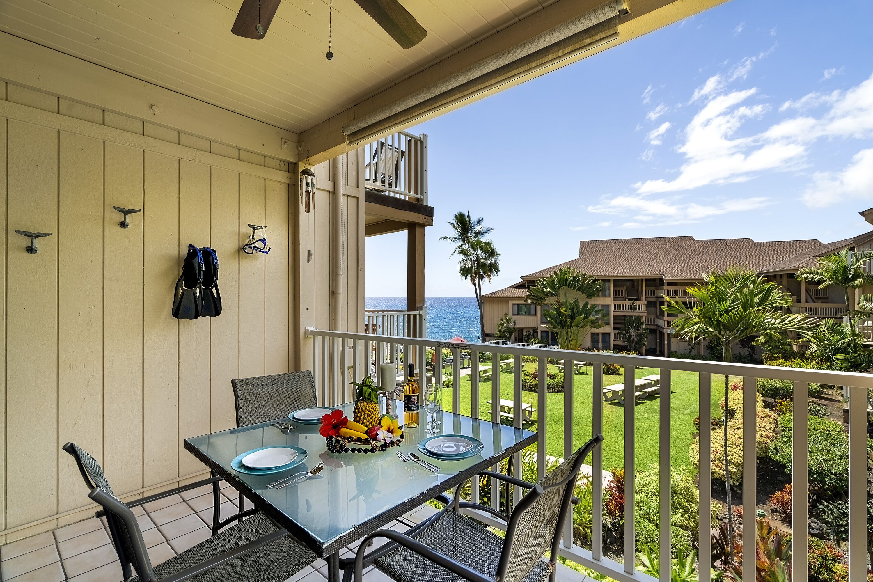 A balcony with a glass table set for dining, overlooking a well-maintained garden and a view of the ocean.