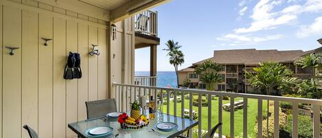 A balcony with a glass table set for dining, overlooking a well-maintained garden and a view of the ocean.