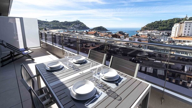 A balcony with a table set for dining, overlooking a coastal town with buildings and hills. - La Concha Terrace - The Rentals Collection (San Sebastián)