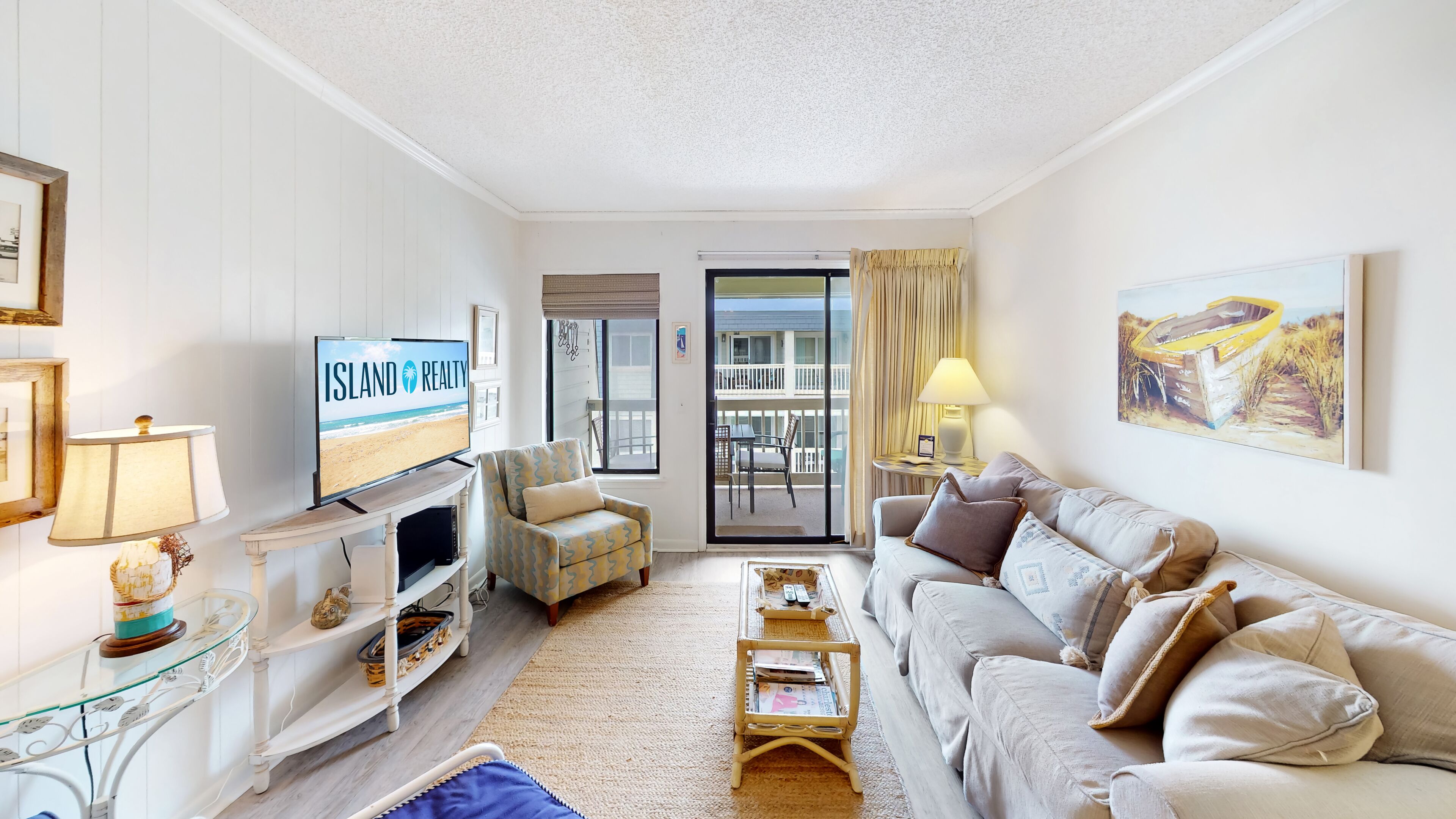 Una sala de estar con un televisor que muestra 'Island Realty', un sofá, una mesa de centro, un mueble para la televisión, una mesa de vidrio con objetos decorativos y un balcón con vista a la playa.