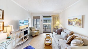 Un salon avec un téléviseur diffusant l’émission « Island Realty », un canapé, une table basse, un meuble pour téléviseur, une table en verre ornée d’objets décoratifs et un balcon avec vue sur la plage.