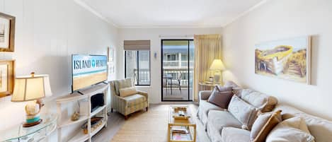A living room with a TV displaying 'Island Realty', a sofa, a coffee table, a TV stand, a glass table with decorative items, and a balcony with a view of the beach.