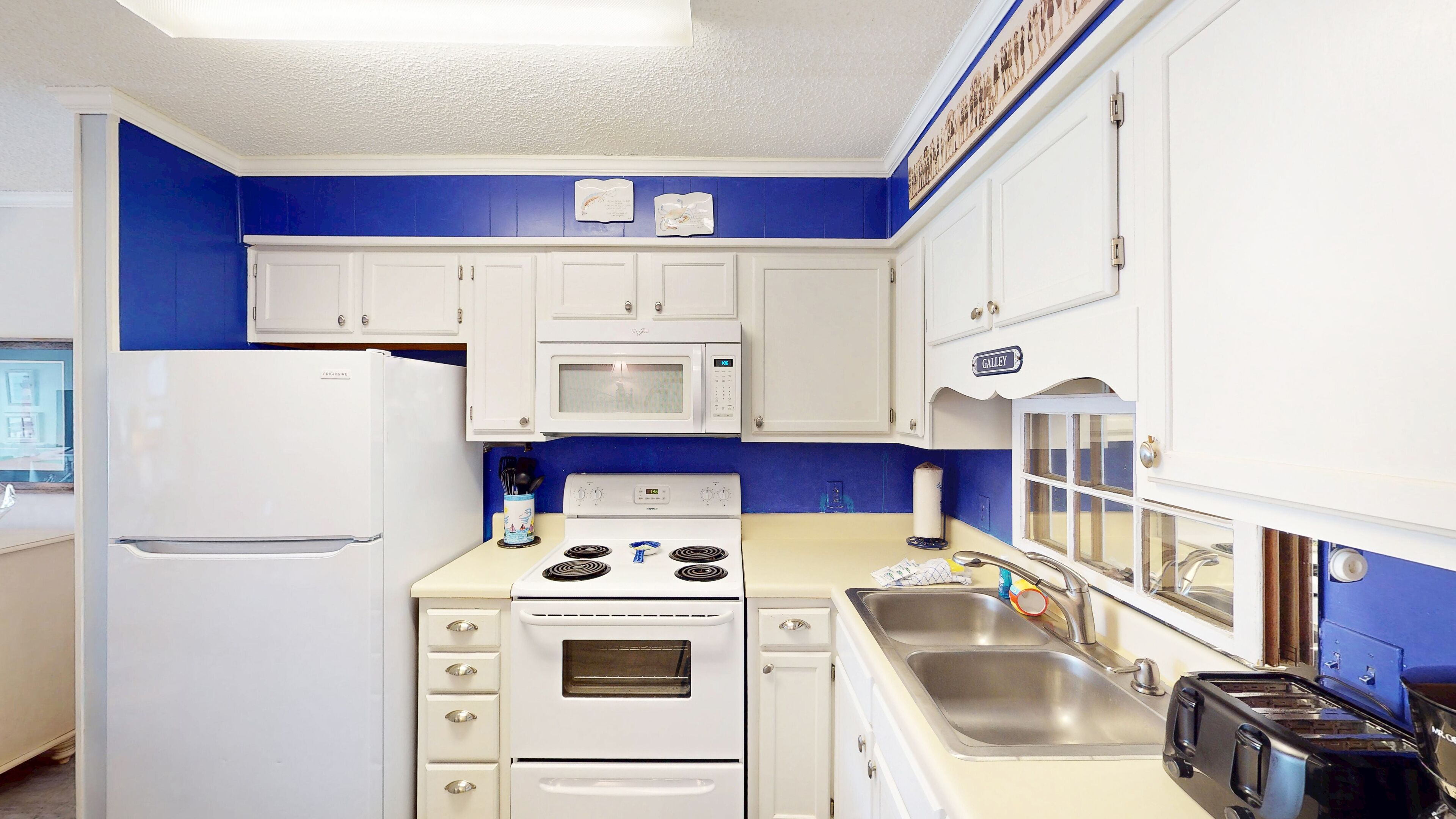 Eine kompakte Küche mit weißen Geräten, darunter ein Kühlschrank, eine Mikrowelle, ein Herd und ein Backofen. Es gibt ein Waschbecken mit Wasserhahn und einen Toaster auf der Arbeitsfläche.