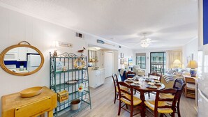 Un coin repas est aménagé avec une table en bois, dressée pour quatre personnes, et entourée de chaises en osier. On aperçoit, sur la gauche, un espace cuisine avec des meubles blancs et un plan de travail bleu.