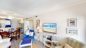 Un salon comprenant un coin repas, un téléviseur diffusant l’émission « ISLAND REALTY », et une décoration murale sur le thème de la plage.