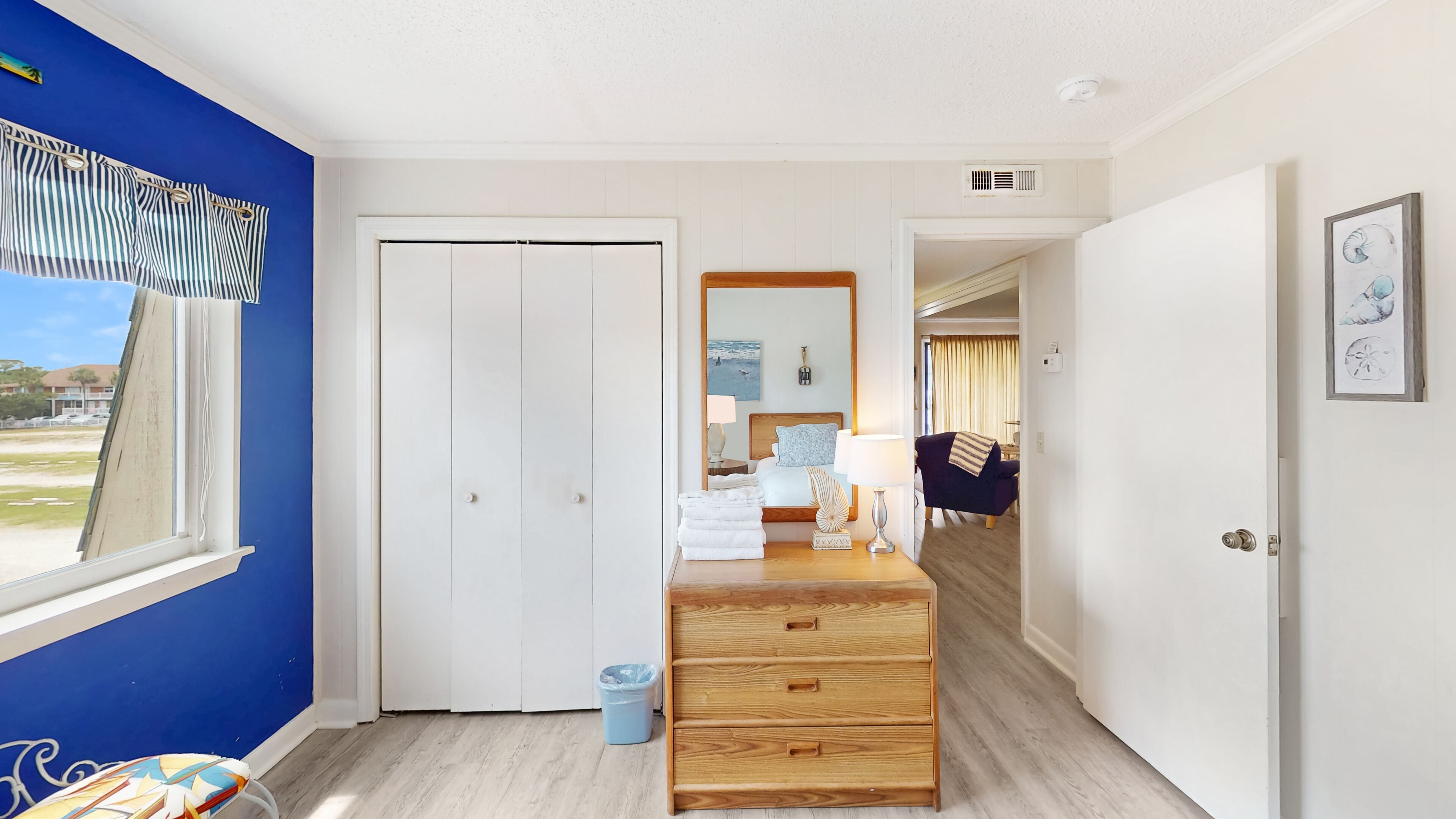 A hotel room with a large window, a wooden dresser, a bed with a headboard, and a view of the beach.