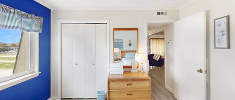 A hotel room with a large window, a wooden dresser, a bed with a headboard, and a view of the beach.