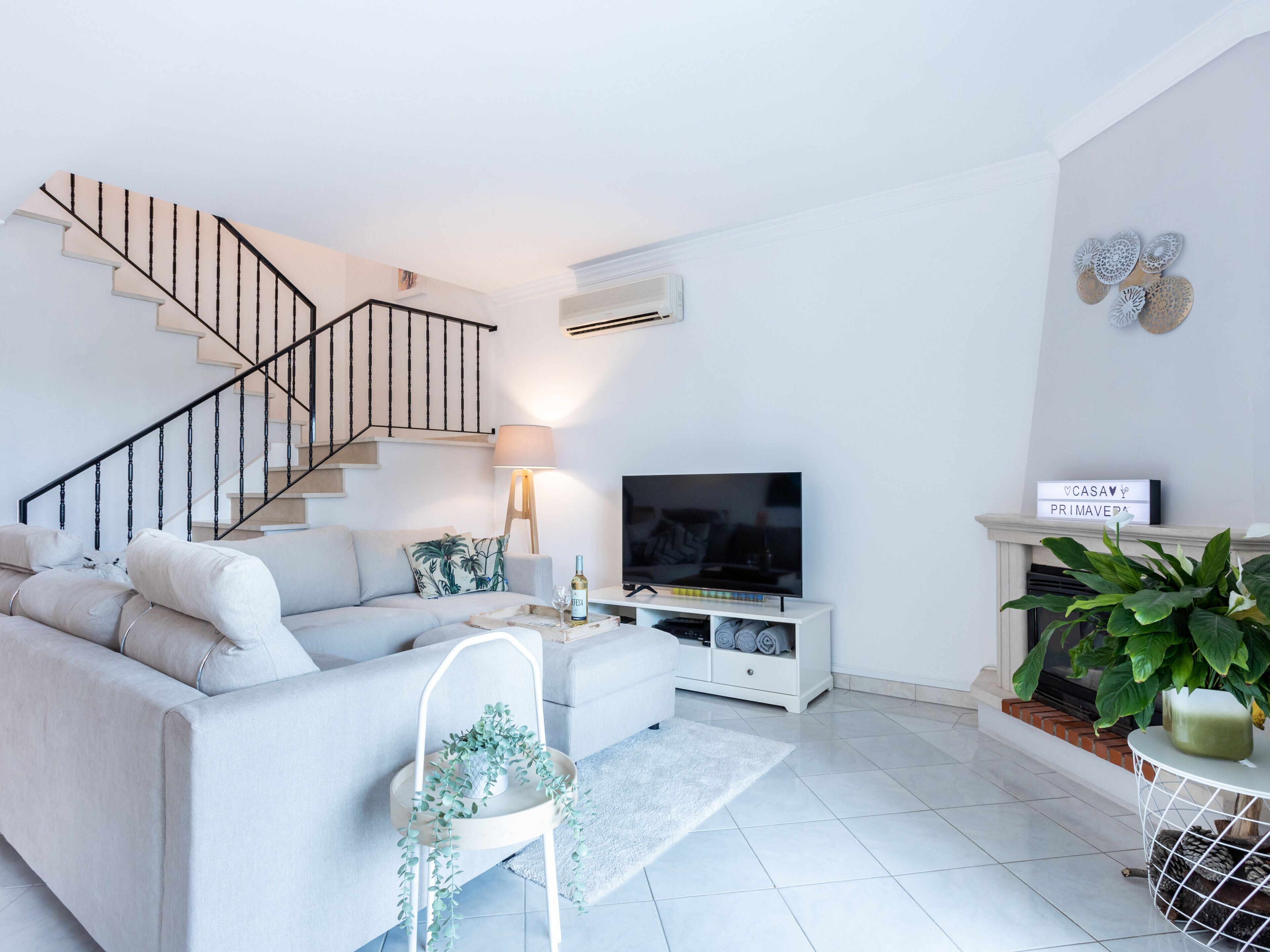 A modern living room with a staircase, a flat-screen TV, a fireplace, and a large potted plant.