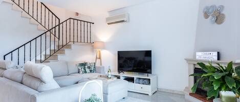 A modern living room with a staircase, a flat-screen TV, a fireplace, and a large potted plant.