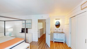 A bedroom with a bed, a desk, a chair, a TV, a bathroom with a sink, mirror, and a framed picture on the wall.