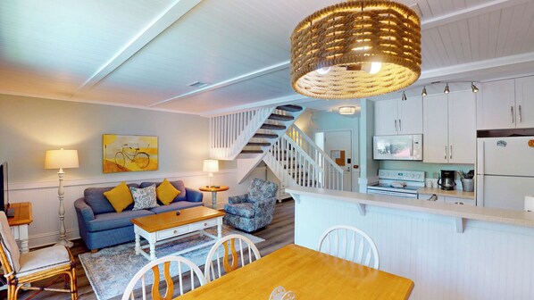 A modern living room with a sofa, coffee table, and a dining area with chairs. There is a staircase leading to another level, and a kitchen area with appliances.