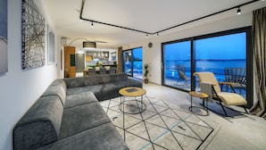 Ein modernes Wohnzimmer mit einer grauen Eckcouch, einem runden Couchtisch und Meerblick durch große Fenster.