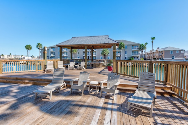 A wooden deck with lounge chairs and a covered seating area, overlooking a body of water and a row of buildings. - Waterfront Retreat | Pool, Hot Tub & Fishing Access (Corpus Christi)