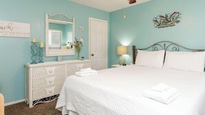 A clean, well-lit bedroom with a white bed, a ceiling fan, a dresser, and a decorative mirror.