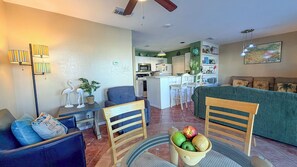 A living room with a sofa, chairs, a dining table with fruit, a kitchen area, and a dining bar.
