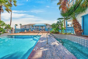 A swimming pool with a blue and white tiled edge, a wooden deck, and a building with a blue roof in the background. - Waterfront Retreat | Pool, Hot Tub & Fishing Access (Corpus Christi)