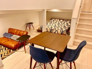 A compact living space with a sofa, a dining table, and two chairs. A staircase is visible in the background.