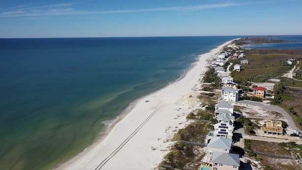 An aerial view of a coastal area with a sandy beach, houses, and a clear view of the ocean.