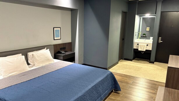 A hotel room with a large bed, a bedside table with a phone, a framed picture on the wall, and a bathroom visible through an open door. - Hotel Le-Gar (Monterrey)