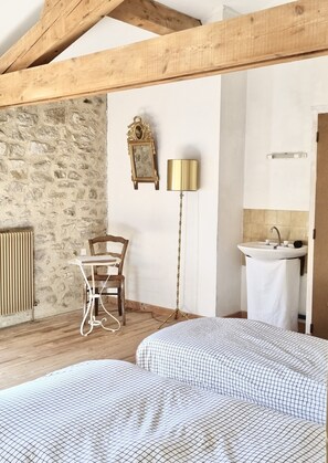 Un dormitorio con techo de madera, pared de piedra, cama con colcha a cuadros, mesita de noche, una silla, lámpara de pie, espejo y baño con lavamanos y toalla.