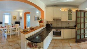 Een moderne keuken met witte kasten, een zwart marmeren aanrechtblad en een ingebouwde oven. Er is een eethoek met witte stoelen en een tafel, en een gang met ingelijste foto’s aan de wand.