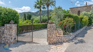 A stone gate with a sign, a gravel path, and a stone wall with a fence and plants. - Finca LHort de Can Bota (Pollensa)