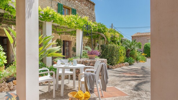 An outdoor patio with white chairs and tables, surrounded by lush greenery and a stone building. - Finca LHort de Can Bota (Pollensa)