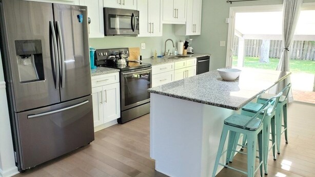 A modern kitchen with white cabinets, stainless steel appliances, a granite countertop, and a small island with bar stools. - Floridays Vacation Home with Pool near to beach by RedAwning (St. Augustine)