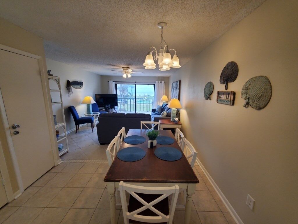 A dining area with a wooden table, white chairs, and blue placemats. There is a ceiling fan above the table and a television in the background.
