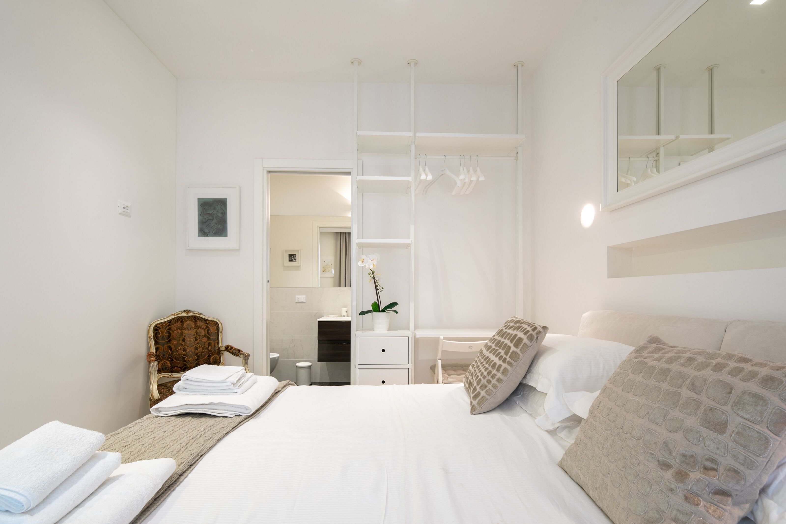 A modern bedroom with a large bed, a chair, a vanity, and a walk-in closet.