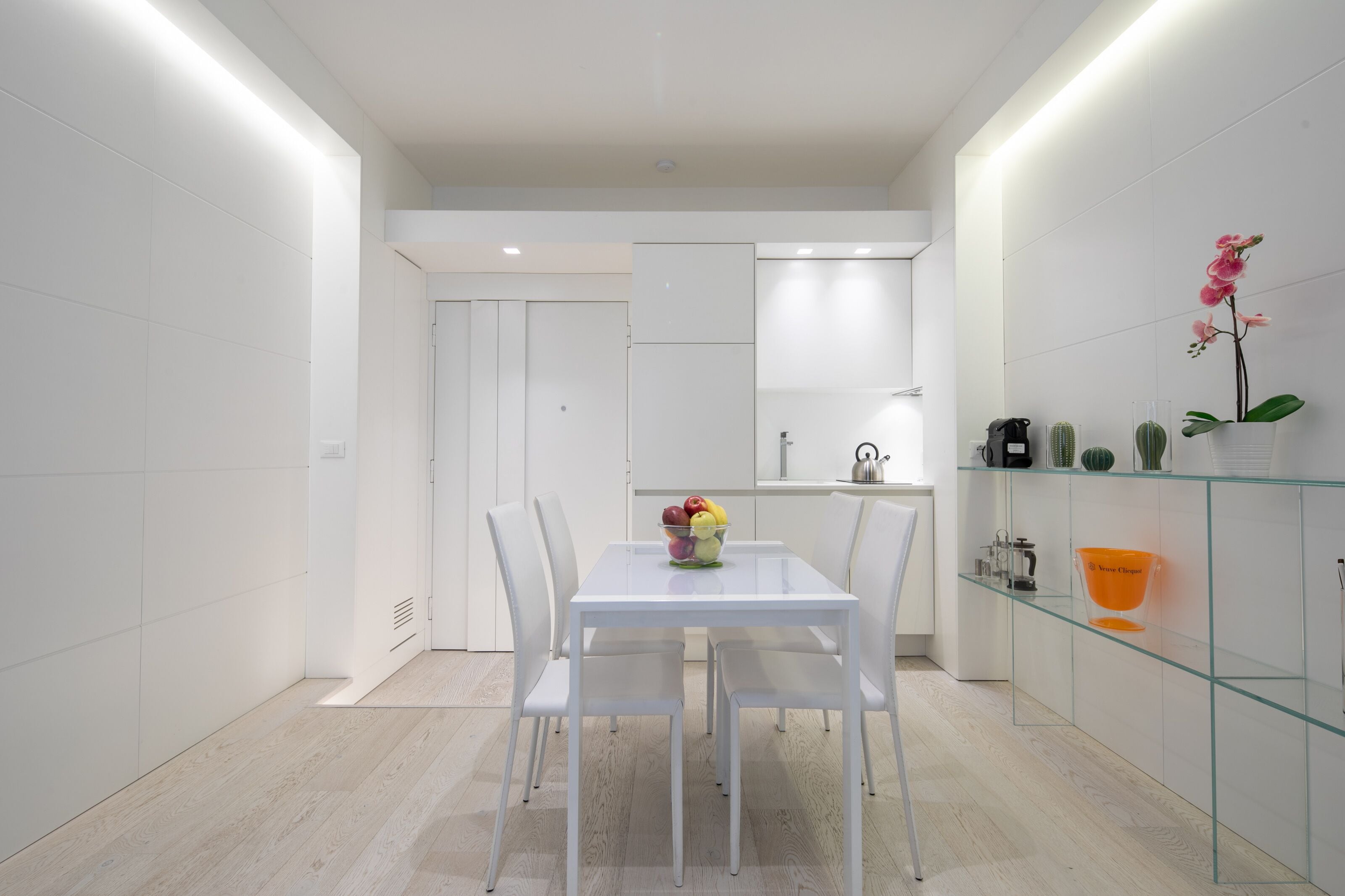 A modern kitchen with white cabinetry, a glass dining table, and chairs. There is a fruit bowl on the table and a cactus on the counter.