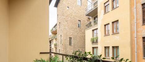 A narrow street with buildings on either side, balconies with plants, and a stone building in the background.