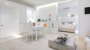 Un salón moderno y minimalista con una mesa de centro de vidrio, un comedor con sillas blancas y un dormitorio con cama y televisión.