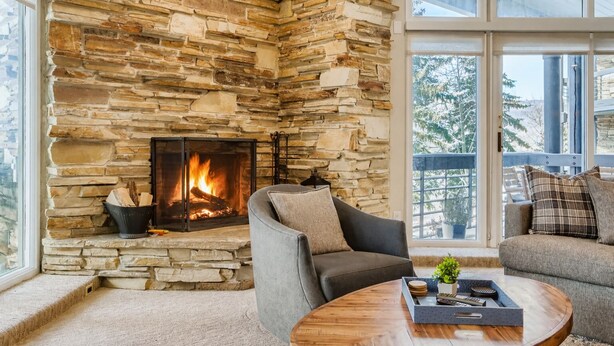 Una chimenea de piedra con fuego, un sillón gris, una mesa de centro de madera y vistas a través de grandes ventanales.