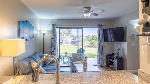 A living room with a sofa, a coffee table, a TV, and a view of the outdoors through a sliding glass door. - Ocean Village Club D15 New 2 Bedroom Condo by RedAwning (St. Augustine)