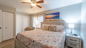 A bedroom with a bed, a ceiling fan, a nightstand, a lamp, a window with curtains, and a painting on the wall. - Ocean Village Club D15 New 2 Bedroom Condo by RedAwning (St. Augustine)