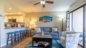 A modern living room with a sofa, coffee table, and bar stools. A kitchen is visible in the background with a microwave and cabinets. - Ocean Village Club D15 New 2 Bedroom Condo by RedAwning (St. Augustine)