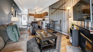 A modern living room with a sofa, coffee table, and chairs. A kitchen with wooden cabinets and a dining area with a table and chairs is visible in the background. - Ten Mile Creek 304 2 Bedroom Condo by RedAwning (Frisco)