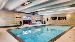 An indoor swimming pool with a tiled floor, a brick wall, and a ceiling with exposed beams. There are lounge chairs and a small seating area with a TV screen. - Ten Mile Creek 304 2 Bedroom Condo by RedAwning (Frisco)